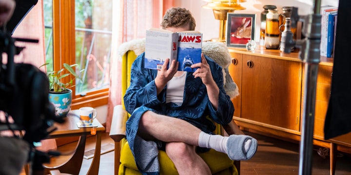 Person relaxing in a cozy living room, wearing a robe and slippers, reading "Jaws" by Peter Benchley next to a window with natural light, a potted plant, and a cup of tea on a side table.